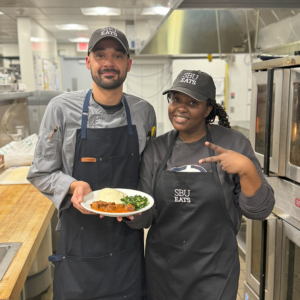 Two chefs posing in a kitchen with a plate of stew, fufu, and greens.