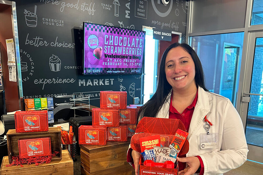 A person holds a red box labeled "The Heart Box," containing snacks, inside a store with an advertisement for chocolate strawberries in the background