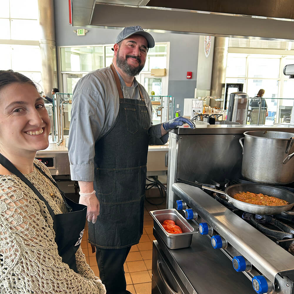 A chef and a woman in a kitchen near a stove with a pan of food.