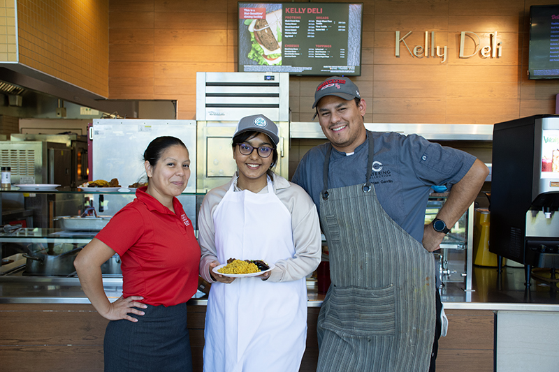 Three kitchen staff members posing together in front of a "Kelly Deli" counter.