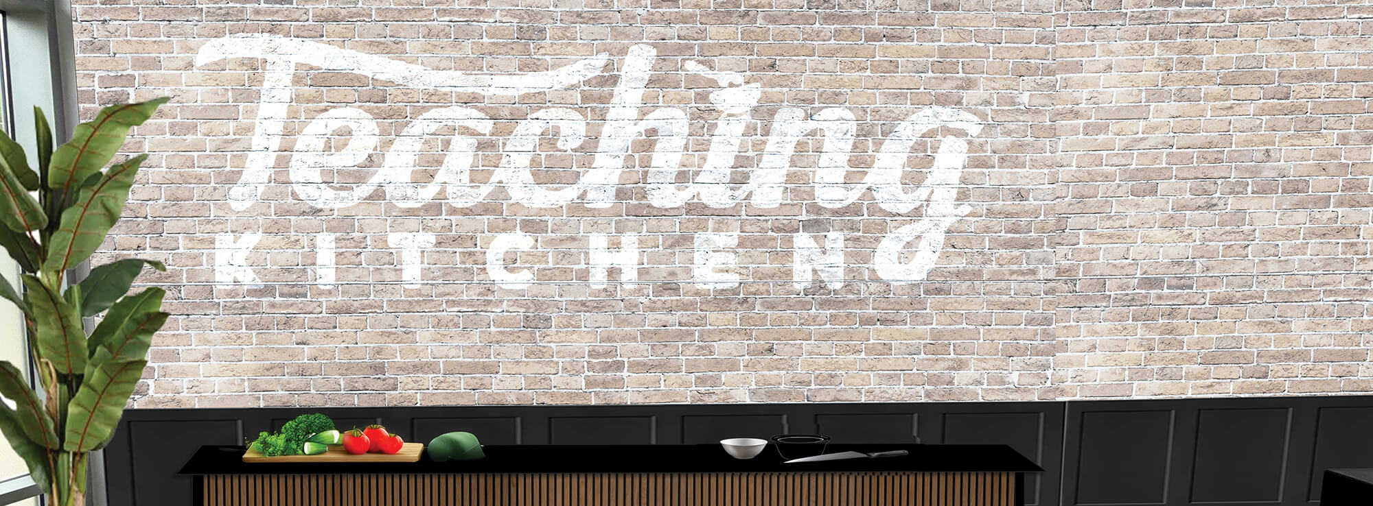 Brick wall with "Teaching Kitchen" in white text, counter with vegetables and kitchenware, and a tall plant on the side