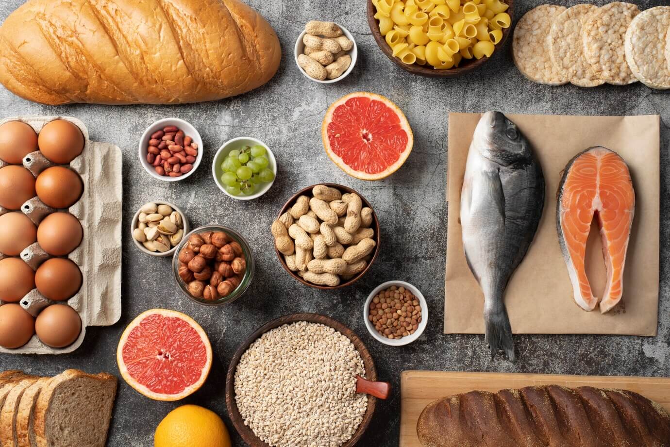 Assorted food items including bread, eggs, pasta, fish, nuts, fruits, and grains are arranged on a gray surface