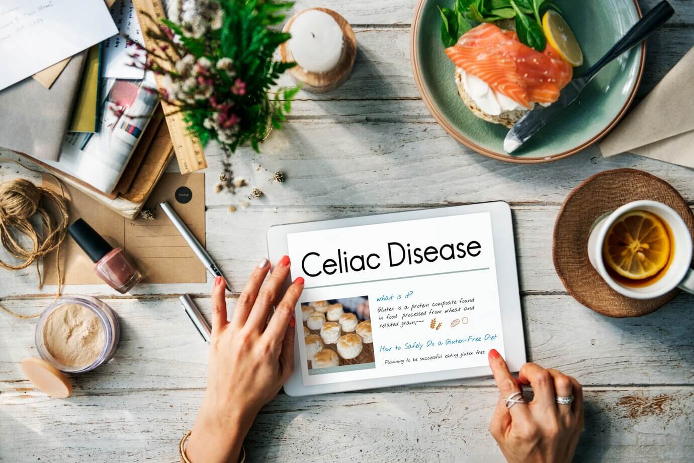 Tablet on a wooden table that reads "Celiac Disease", with a plate of smoked salmon bagel and assorted stationery items