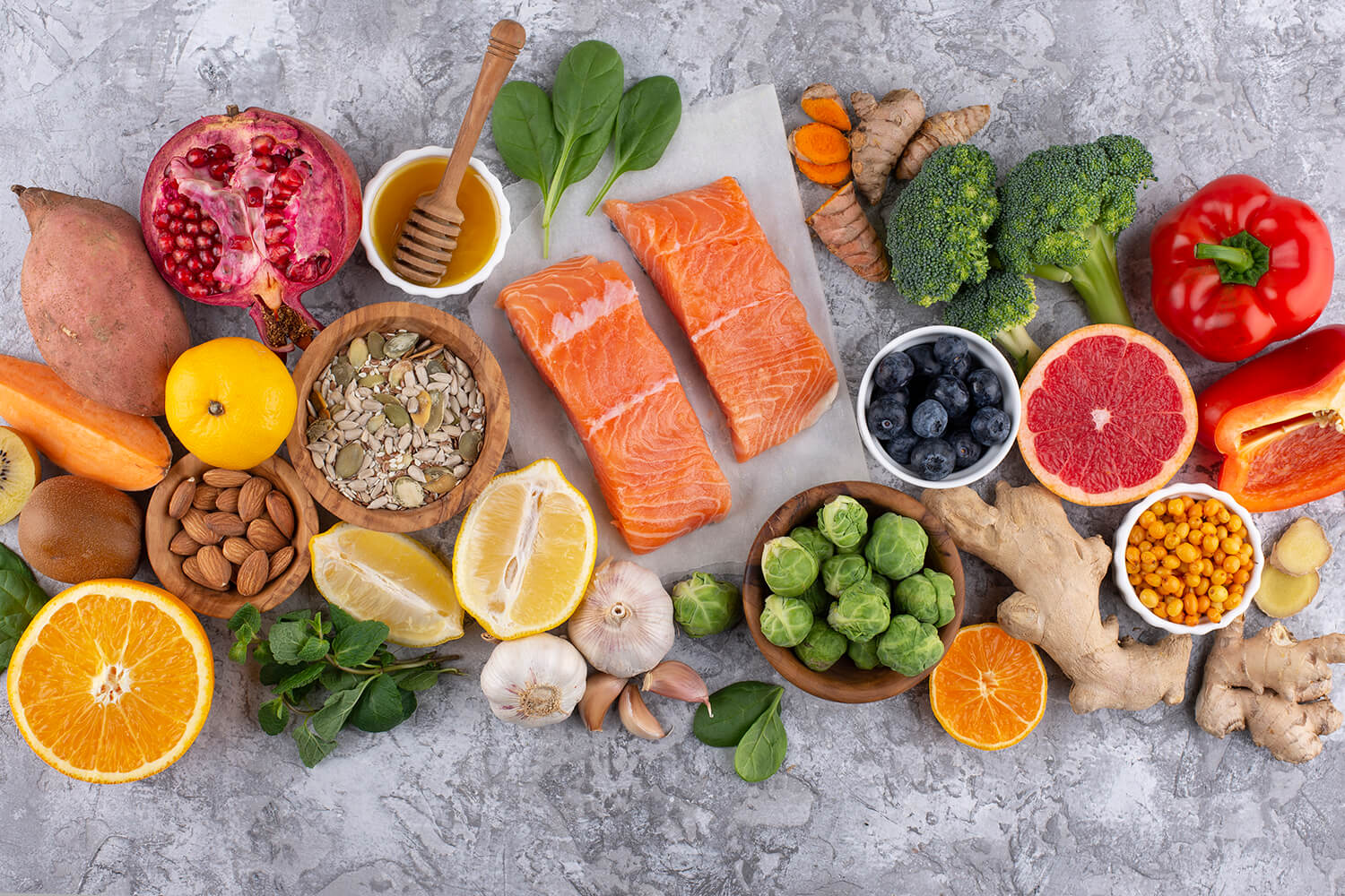 Assorted fresh foods including salmon, fruits, vegetables, nuts, and seeds on a gray surface