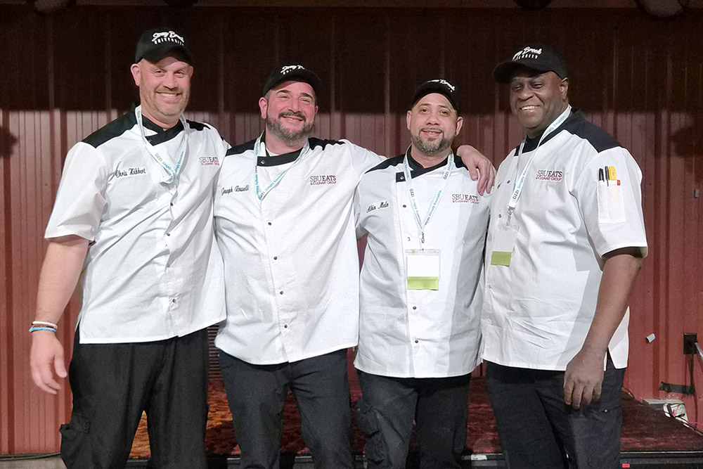 Four men wearing white chef jackets and black caps, standing side by side, smiling