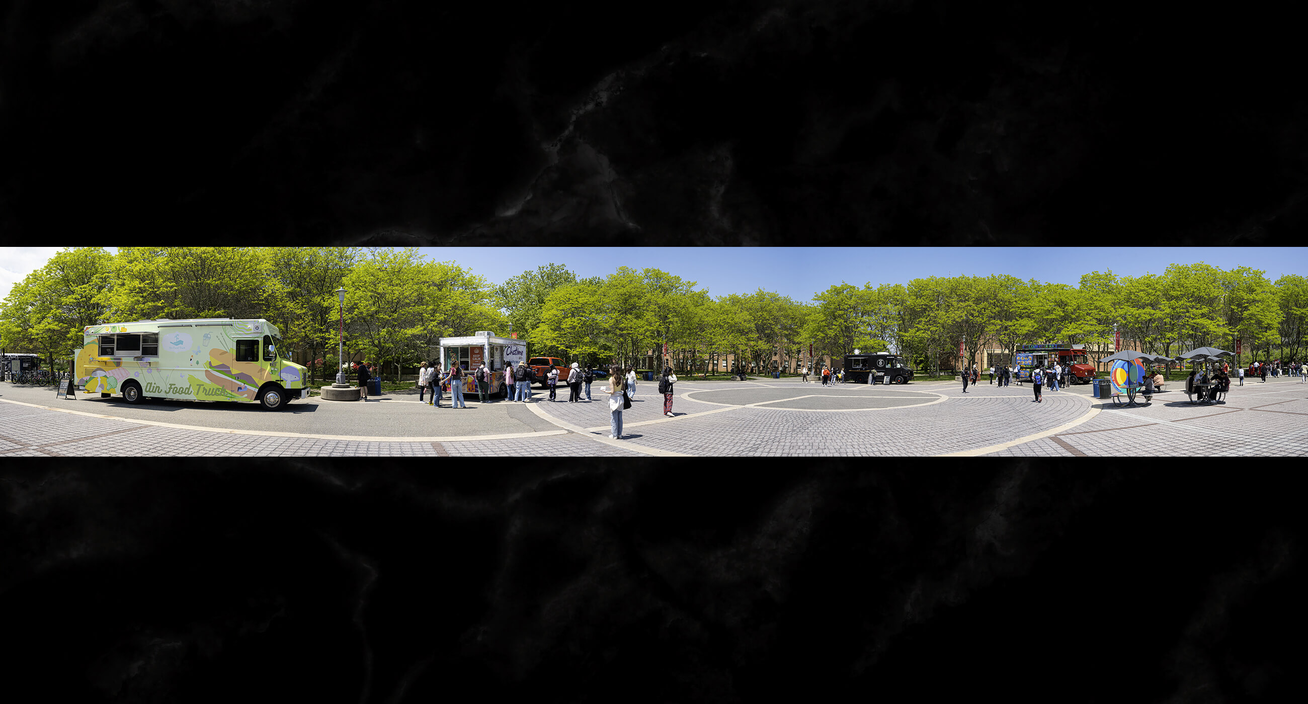 Several food trucks and a crowd of people enjoying the atmosphere in a paved area surrounded by trees