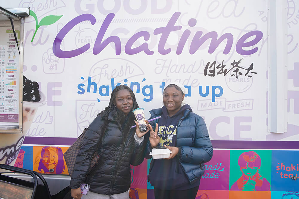 Two young women hold drinks and smile in front of a colorful "Chatime" food truck with vibrant graphics and text.