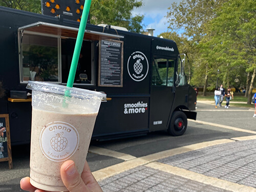 A person holds a smoothie cup from a black food truck labeled "anona," parked in a sunny outdoor location with people and trees in the background