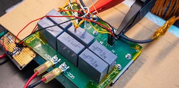 A close-up of a green printed circuit board (PCB) featuring several black relay components mounted in a row, with multiple colored wires soldered and connected across the board.