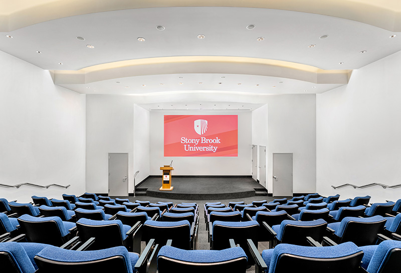 Lecture hall at Stony Brook University with blue seating, a podium, and a large screen displaying the university logo, under soft white lighting.
