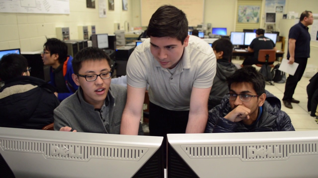 A group of students working together at desktop computers in a classroom. One student stands and leans over the desk, pointing at the screen while two seated students look on attentively. Other students and computers are visible in the background, suggesting a collaborative lab or class setting.