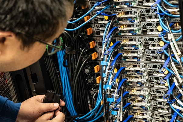 A person inspects a server rack, filled with tangled blue and black cables and various network devices. The setting is technical and focused.