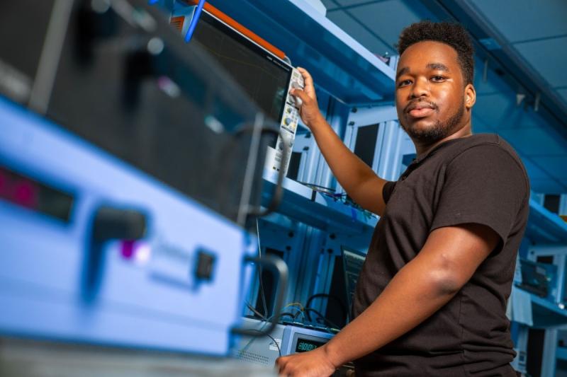 A person in a black shirt stands in a tech lab, confidently adjusting equipment on a shelf. The setting conveys focus and expertise in a professional environment.