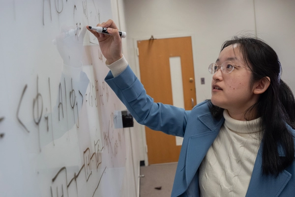 Woman in a blue blazer writing an equation on a whiteboard.