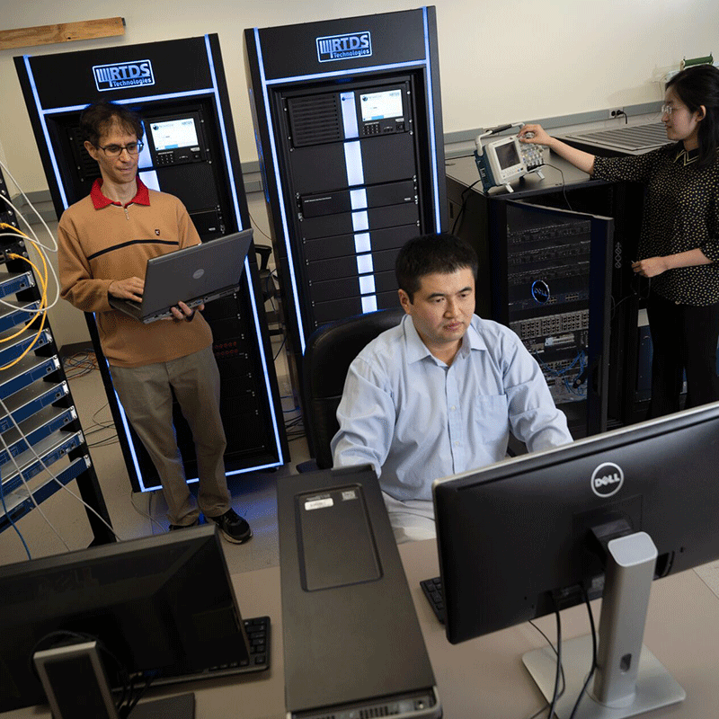 Three colleagues in a lab work with computers and RTDS equipment; one at a desk, another with a laptop, and a third adjusting a device for the "AI-Grid" they have developed.