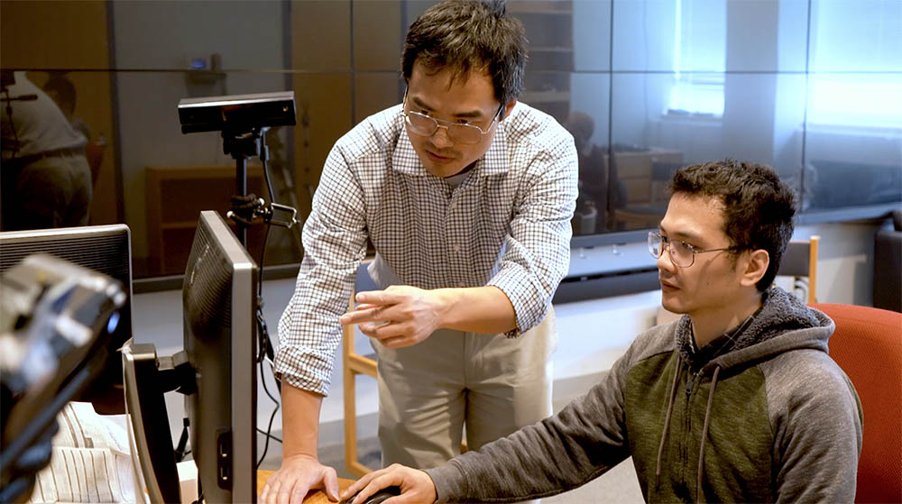 A man in a checked shirt instructs another man seated at a computer. The setting is a modern, well-lit office. Both appear focused and engaged.