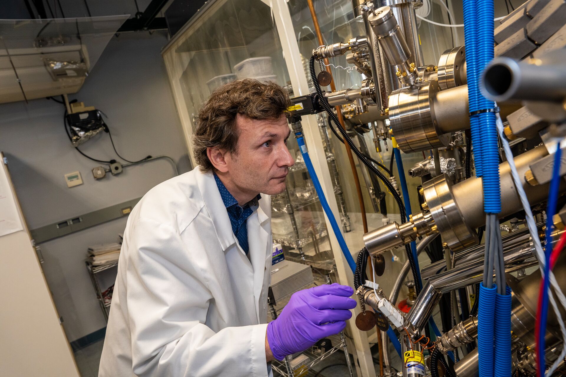 A professor in a lab coat and purple gloves examines complex scientific equipment in a laboratory setting.