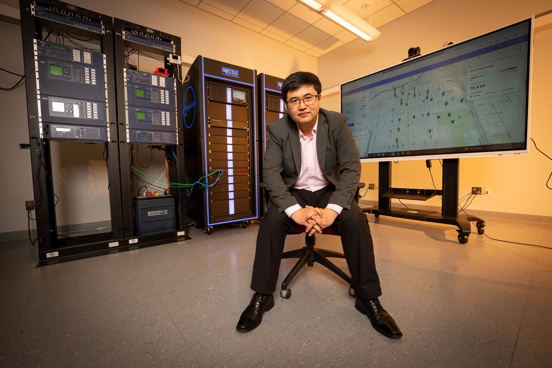 A professor in a suit sits on a chair in a modern lab with AI server racks and a large display showing a map on the screen.