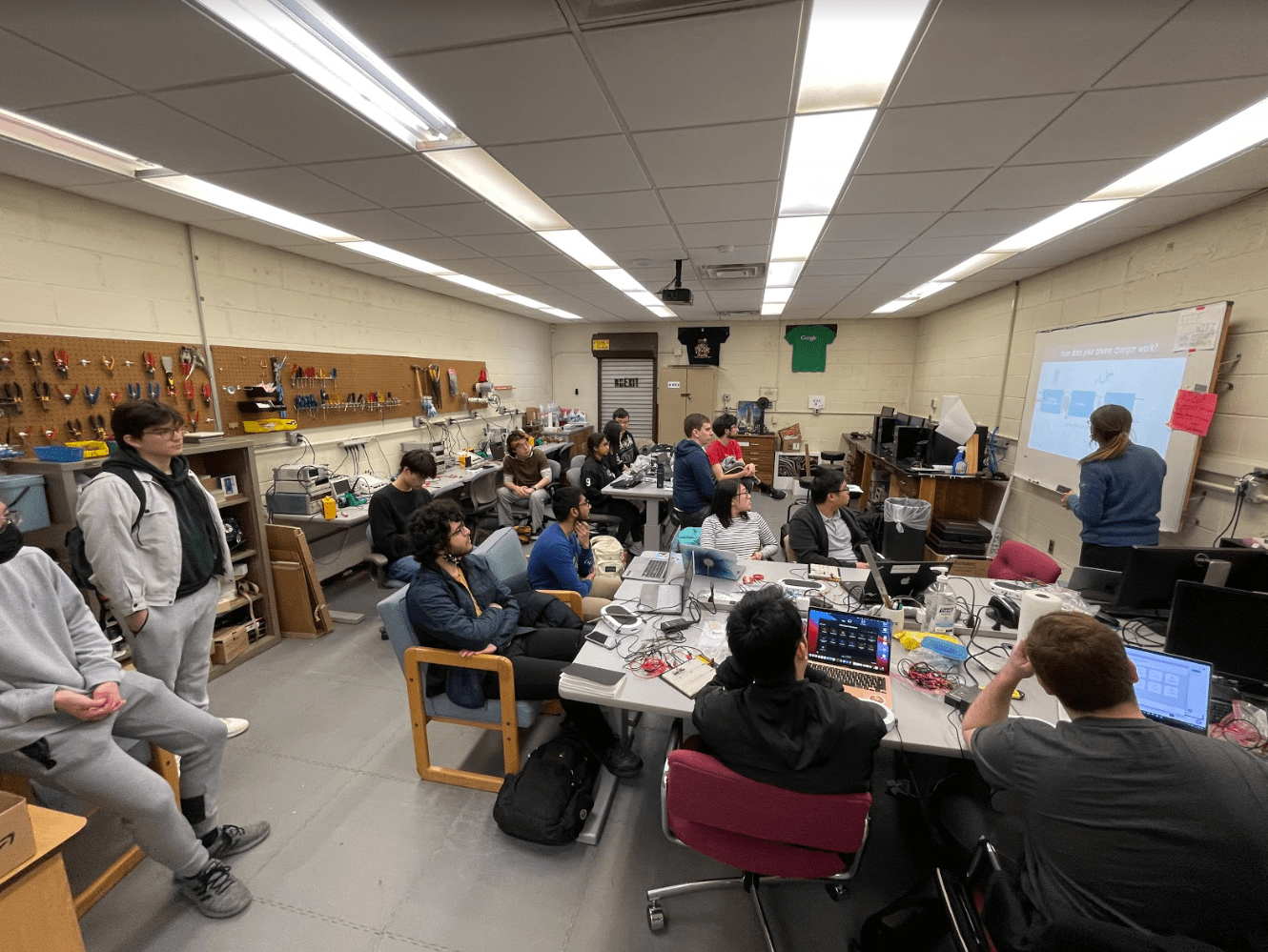A room filled with students attentively watching a presentation on a screen. Laptops and electronic equipment are on the tables, creating an engaging learning environment.