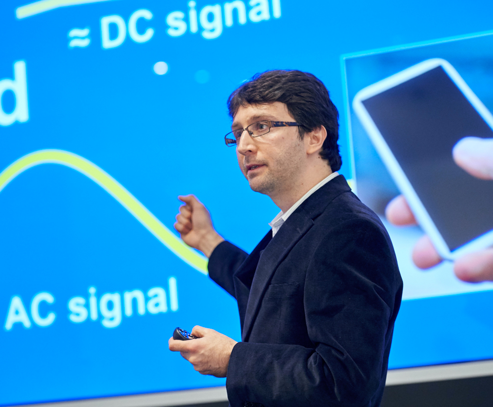 Person in a dark blazer gives a presentation on AC and DC signals, pointing to a graph on a blue screen. He appears focused and analytical.
