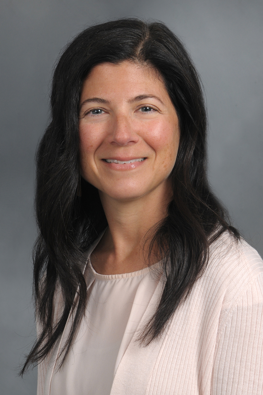 Caucasian woman with long, dark brown side parted wavy hair, wearing a ballerina pink shirt and white cardigan