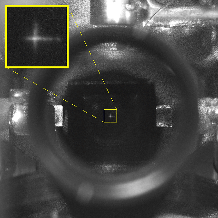 A black-and-white close-up photo of an optical laboratory apparatus, showing a small bright dot inside a central view. Dashed yellow lines point to an inset in the top-left corner, providing a magnified view of the dot as a luminous cross-shaped starburst.