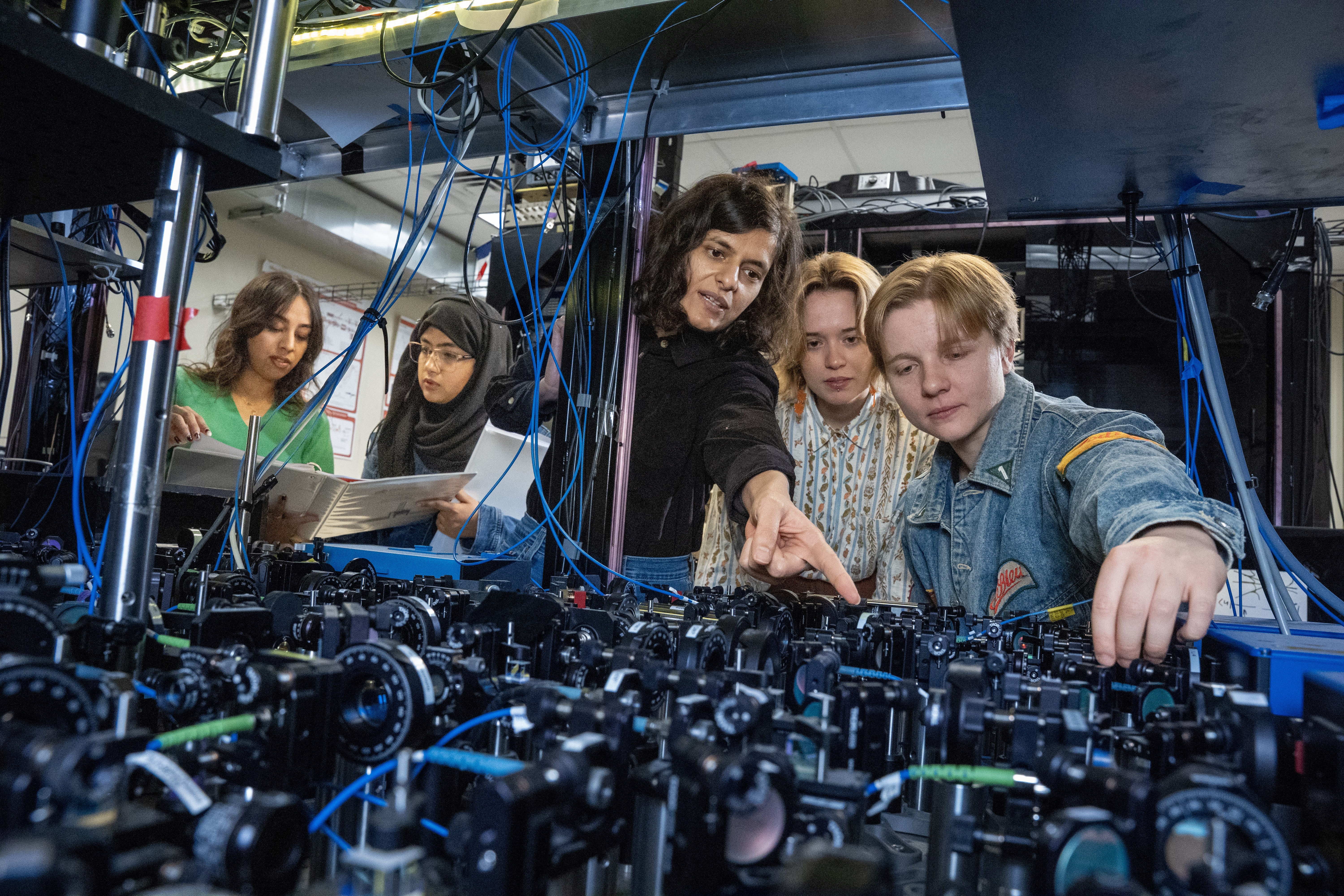 Five researchers collaborating around an optical table with lenses, mirrors, and cables in a physics laboratory.