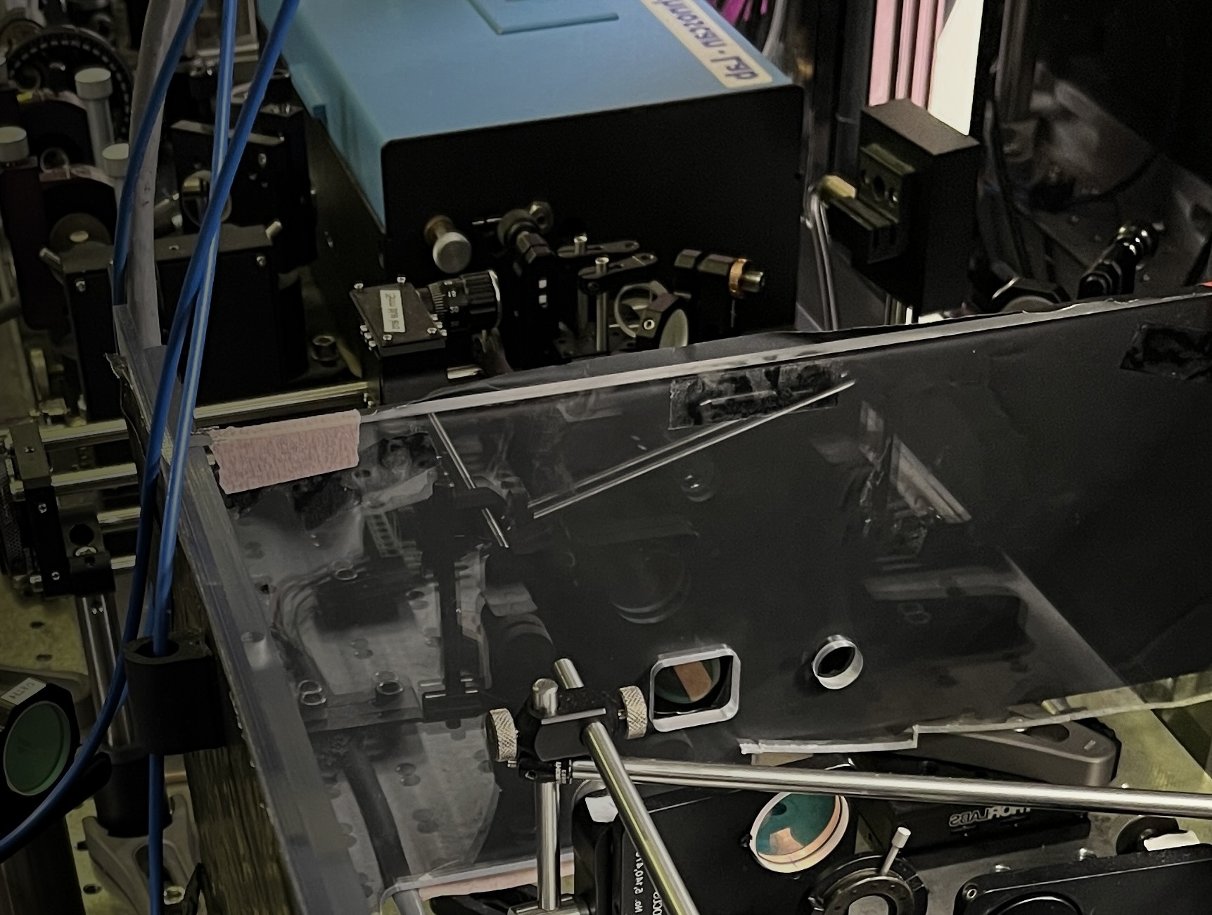 A complex physics laboratory setup featuring a large blue laser housing and optical equipment partially enclosed by a clear plastic protective shield.