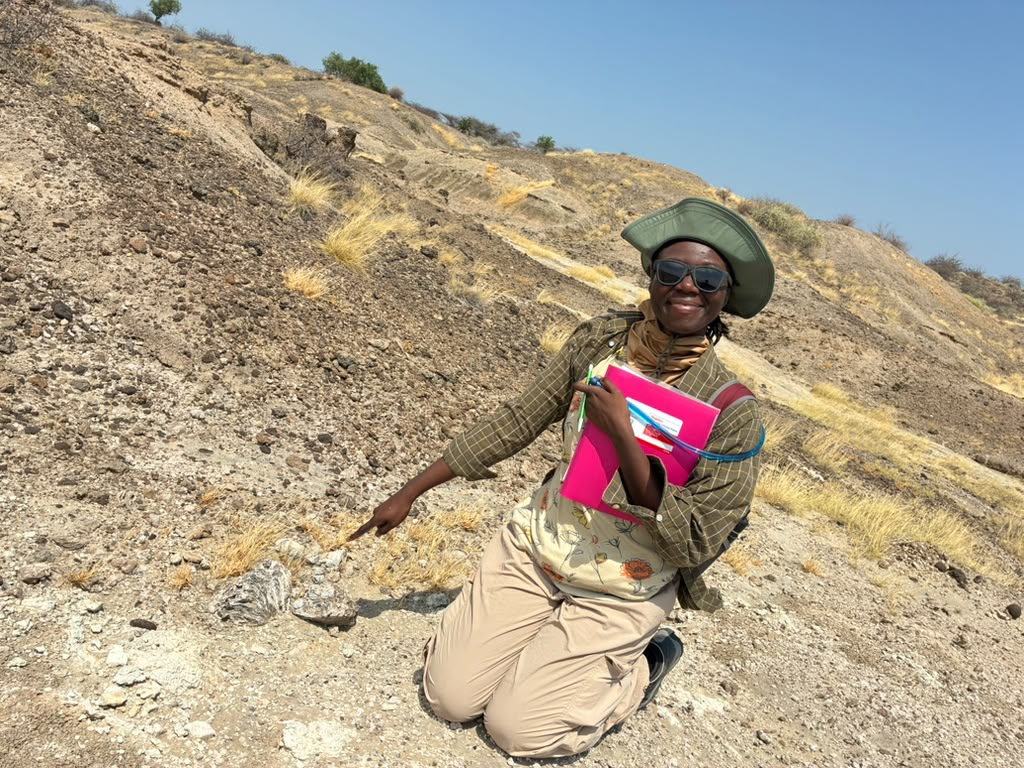 Roheyatou Ceesay sitting in field pointing at fossil