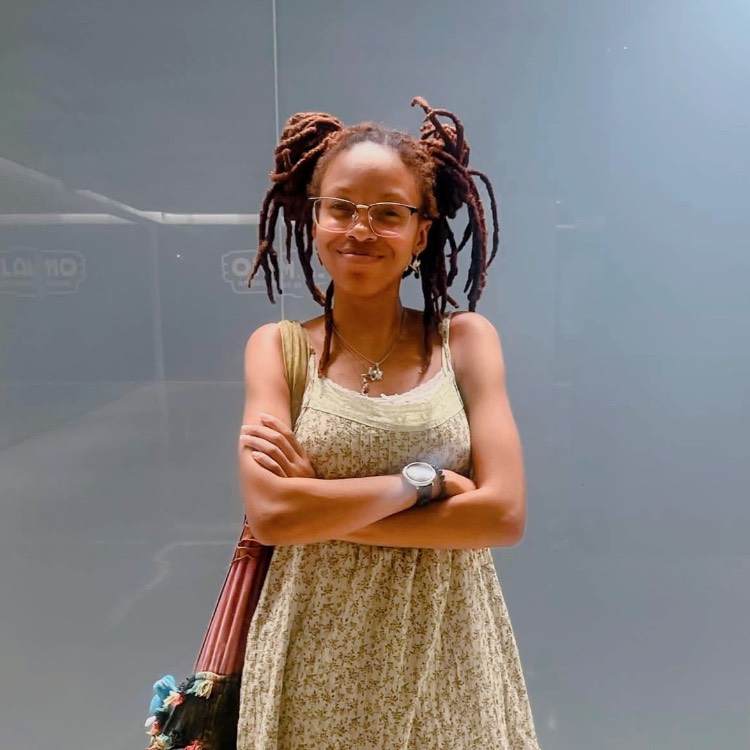 Sheena Luke Girl in a tan sleeveless dress and two messy buns standing with arms crossed