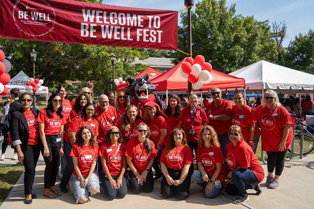 A group of people and Wolfie in red Be Well shirts outside.