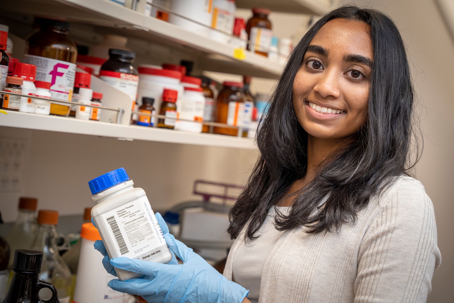A woman smiling and holding a medication bottle. There are shelves with other various medicines behind her.