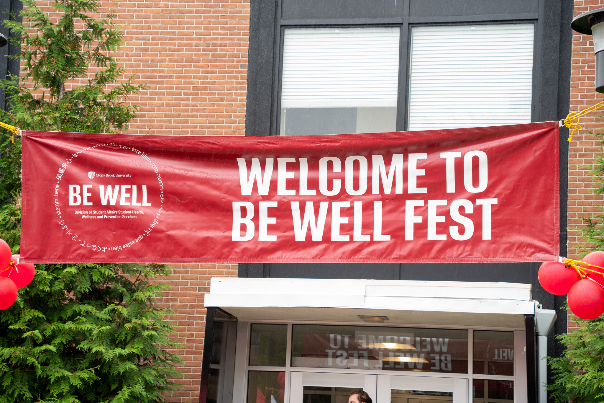 A red sign with white text that says "Welcome to Be Well Fest" hanging in front of a building.