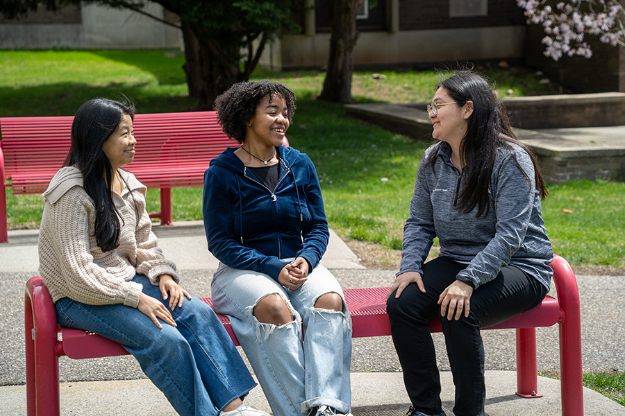 Three students wearing sweaters and jeans talking and sitting on a red bend outside on a sunny spring day.