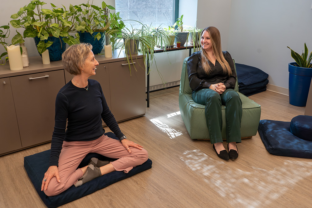 A women sitting cross-legged on a floor mat and a women sitting in a chair. They are in a well-lit room with plants.