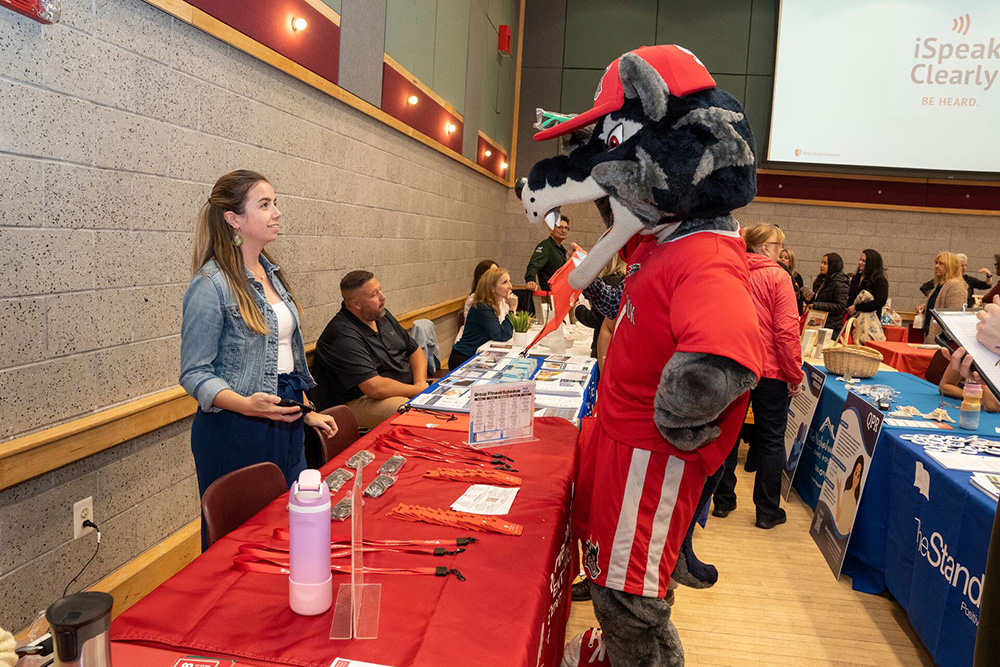 Wolfie standing at a red table across from a woman in an auditorium.