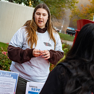 A woman in a grey sweater behind a table talking to a student with black hair outside.