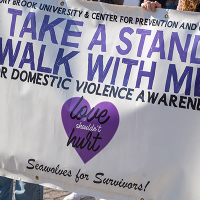 People holding a white sign with blue text that says "TAKE A STAND WALK WITH ME FOR DOMESTIC VIOLENCE AWARENESS".