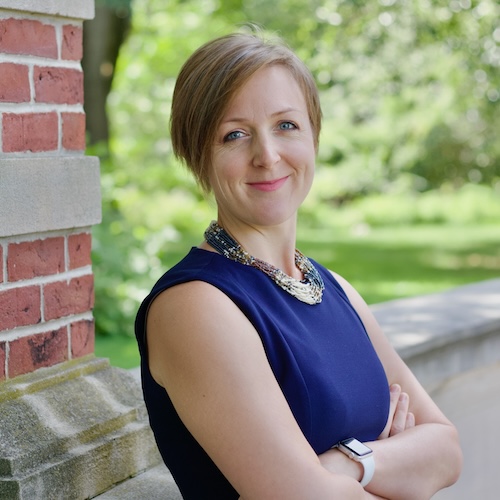 Allison Smagin crossing her arms and smiling outside a brick building surrounded by greenery