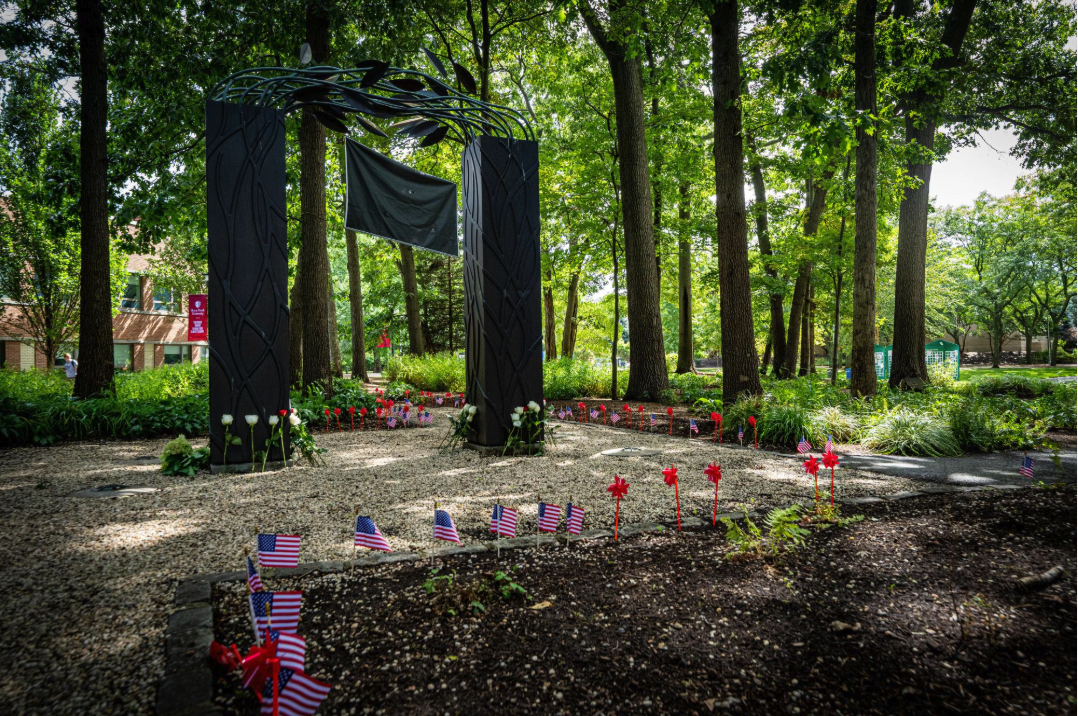 A black archway with leaf motifs and flowers beneath, set among tall trees, small American flags, and red wind spinners on a gravel path.