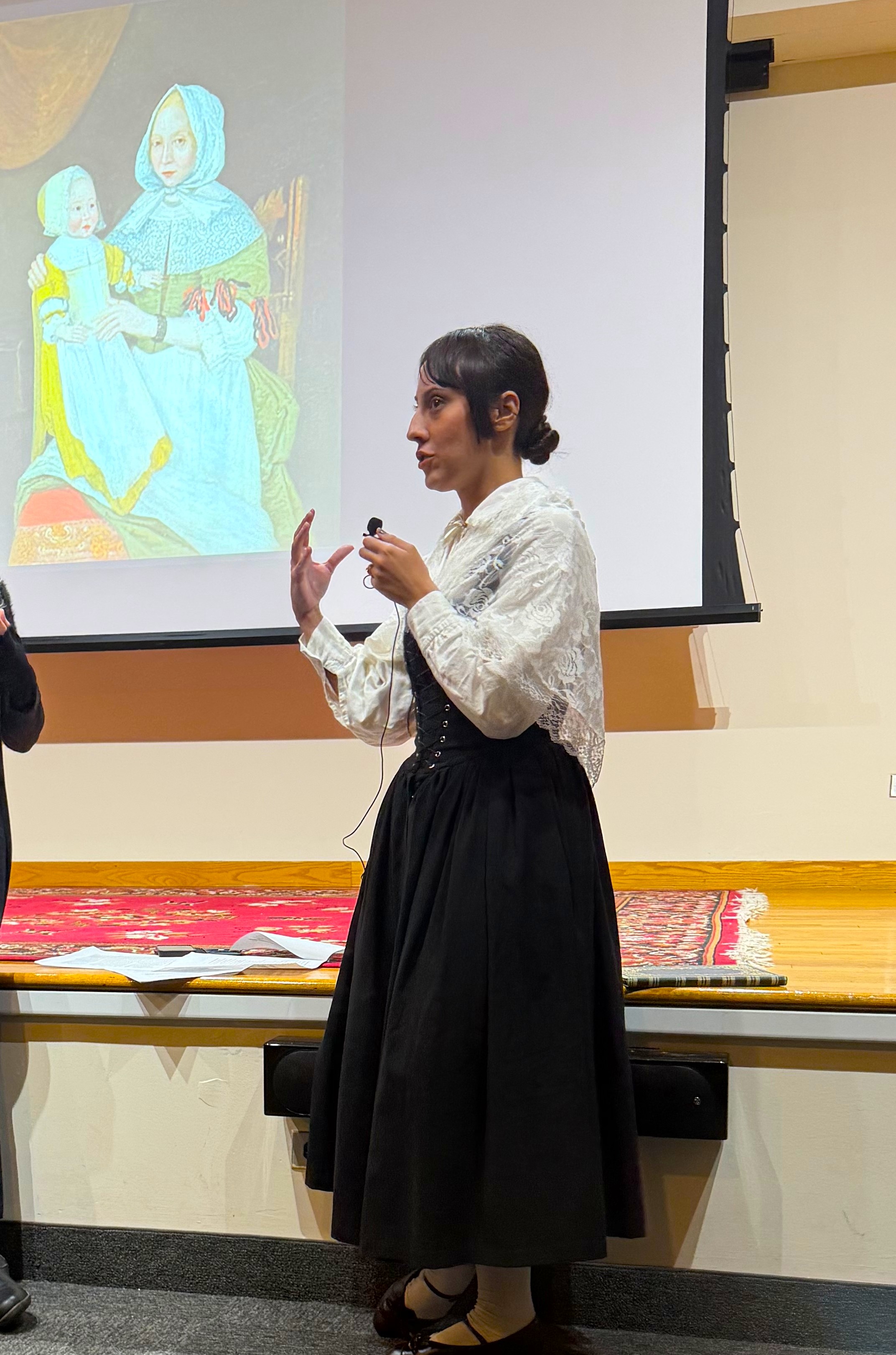 Woman in historical attire speaking on stage with a historical painting projected behind her.