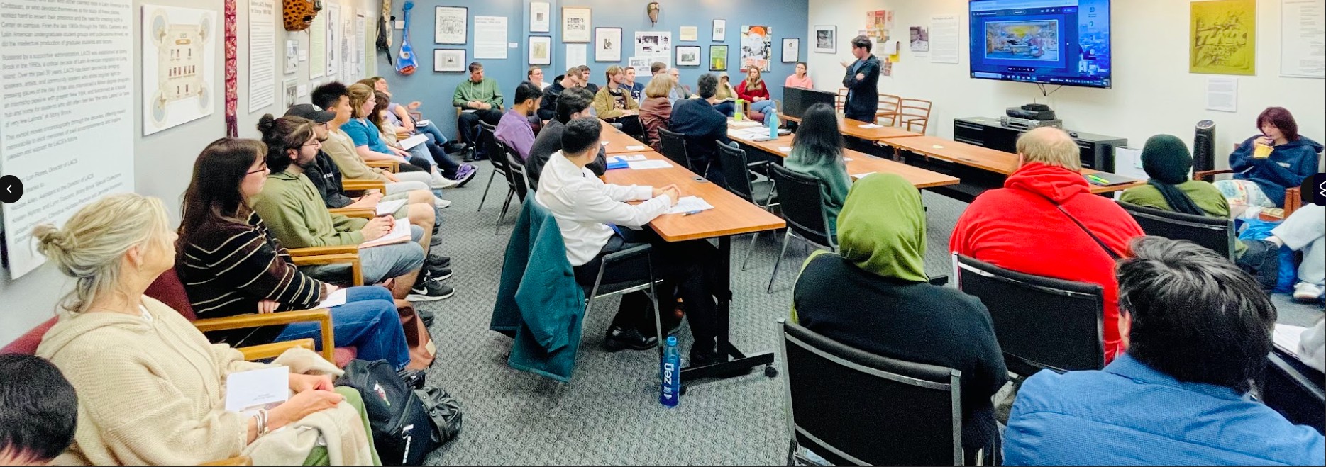 Classroom with people seated around tables, listening to a presentation.
