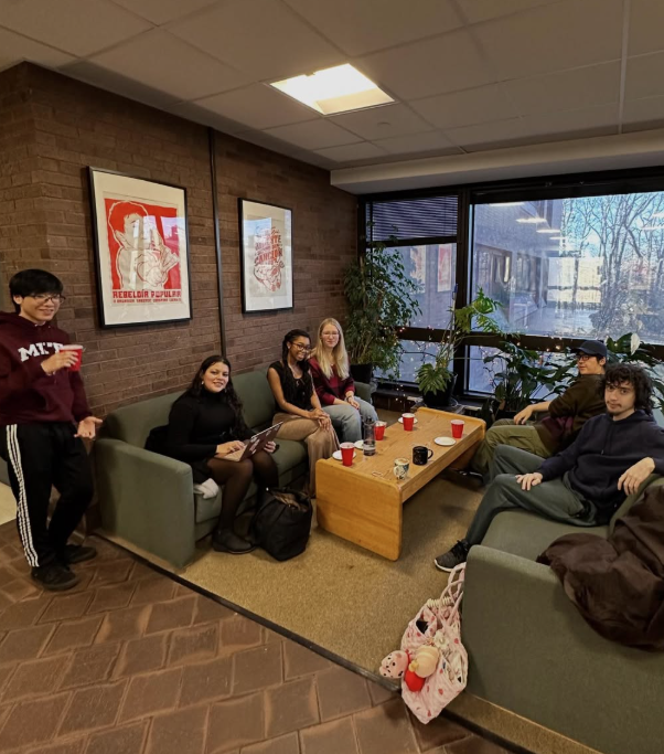 People gathered in a lounge with a wooden coffee table, red cups, and framed posters on a brick wall.
