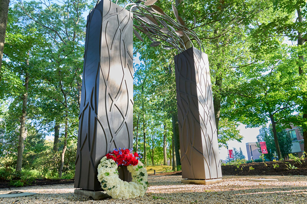 A black archway with leaf motifs and flowers beneath, set among tall trees, small American flags, and red wind spinners on a gravel path.
