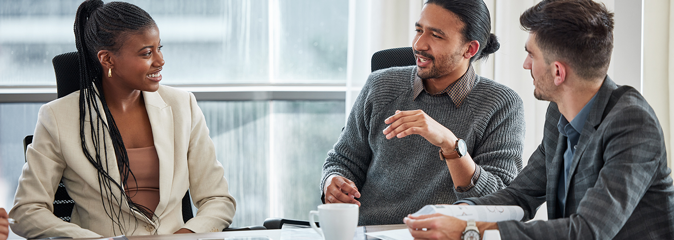 Three professionals engage in a collaborative discussion around a table in a modern, sunlit office with city views in the background.