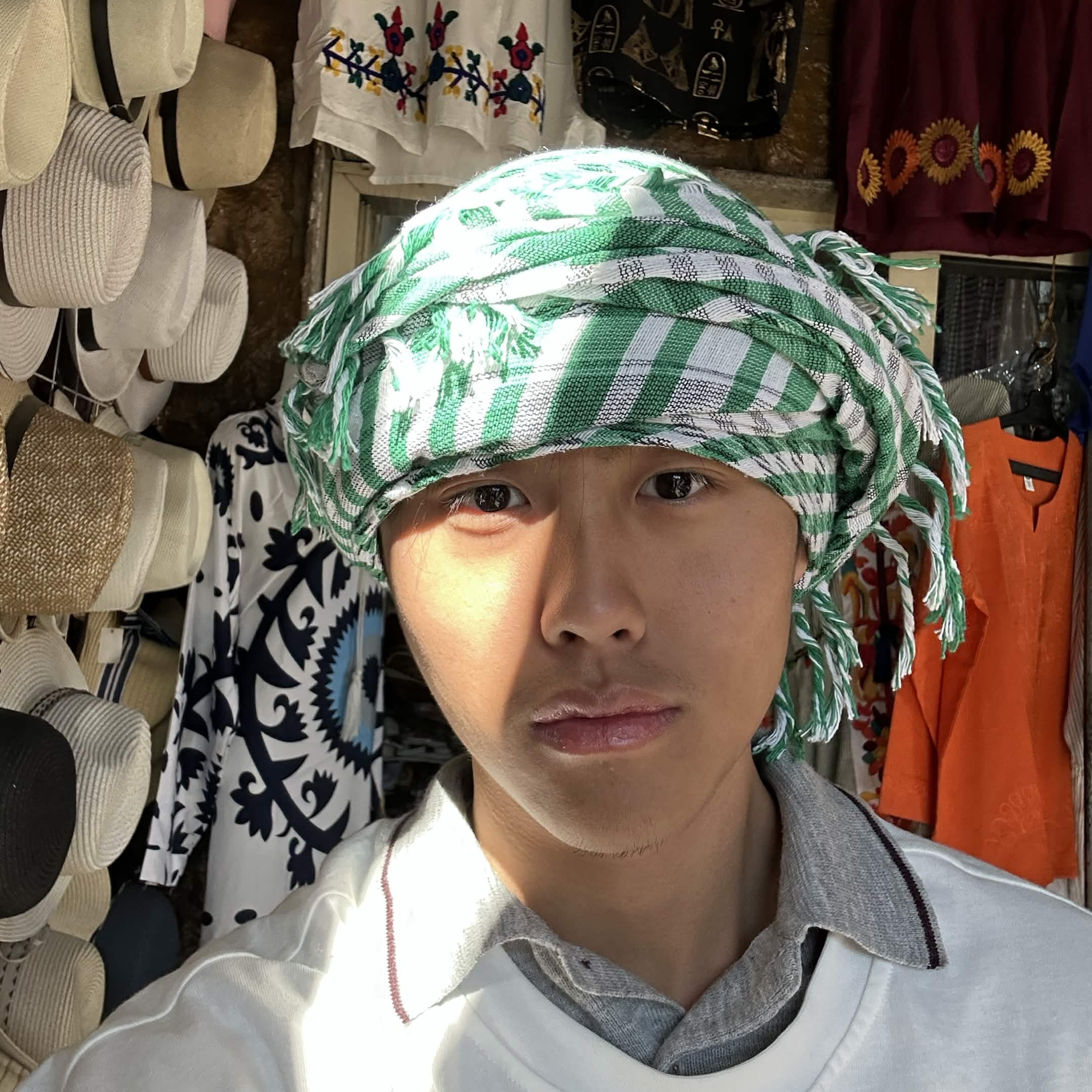 Boy in green and white headscarf and white shirt with local market behind him