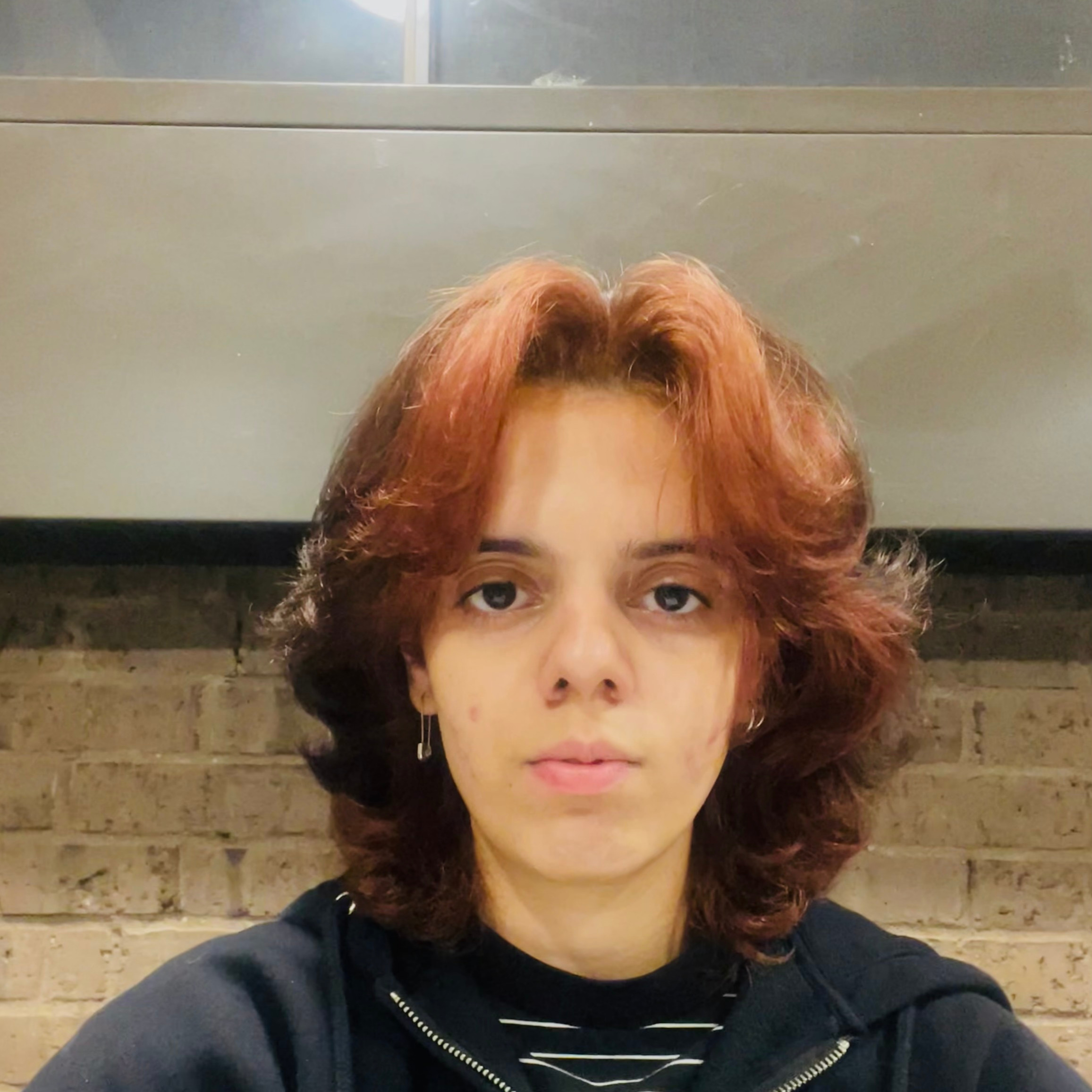 Girl with red and black hair and blue zip up jacket sits in front of a brick wall