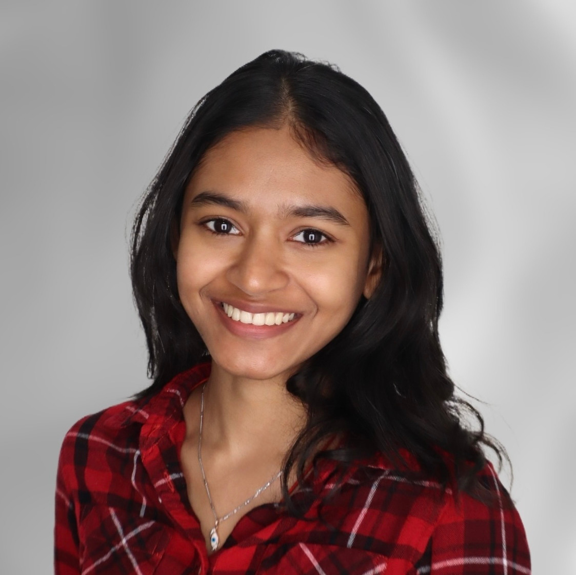Girl with dark hair in a red and black plaid button up smiles up at the camera with white wall behind her