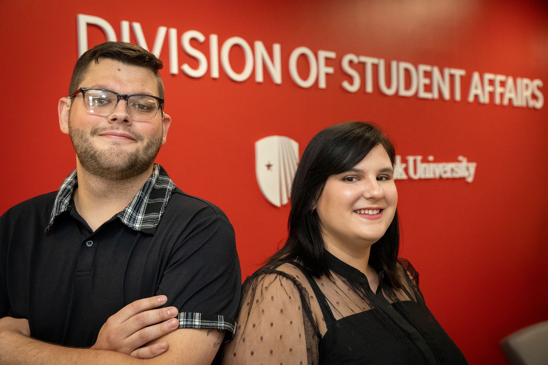 Two smiling professionals pose in front of a red wall reading "Division of Student Affairs" at Stony Brook University.