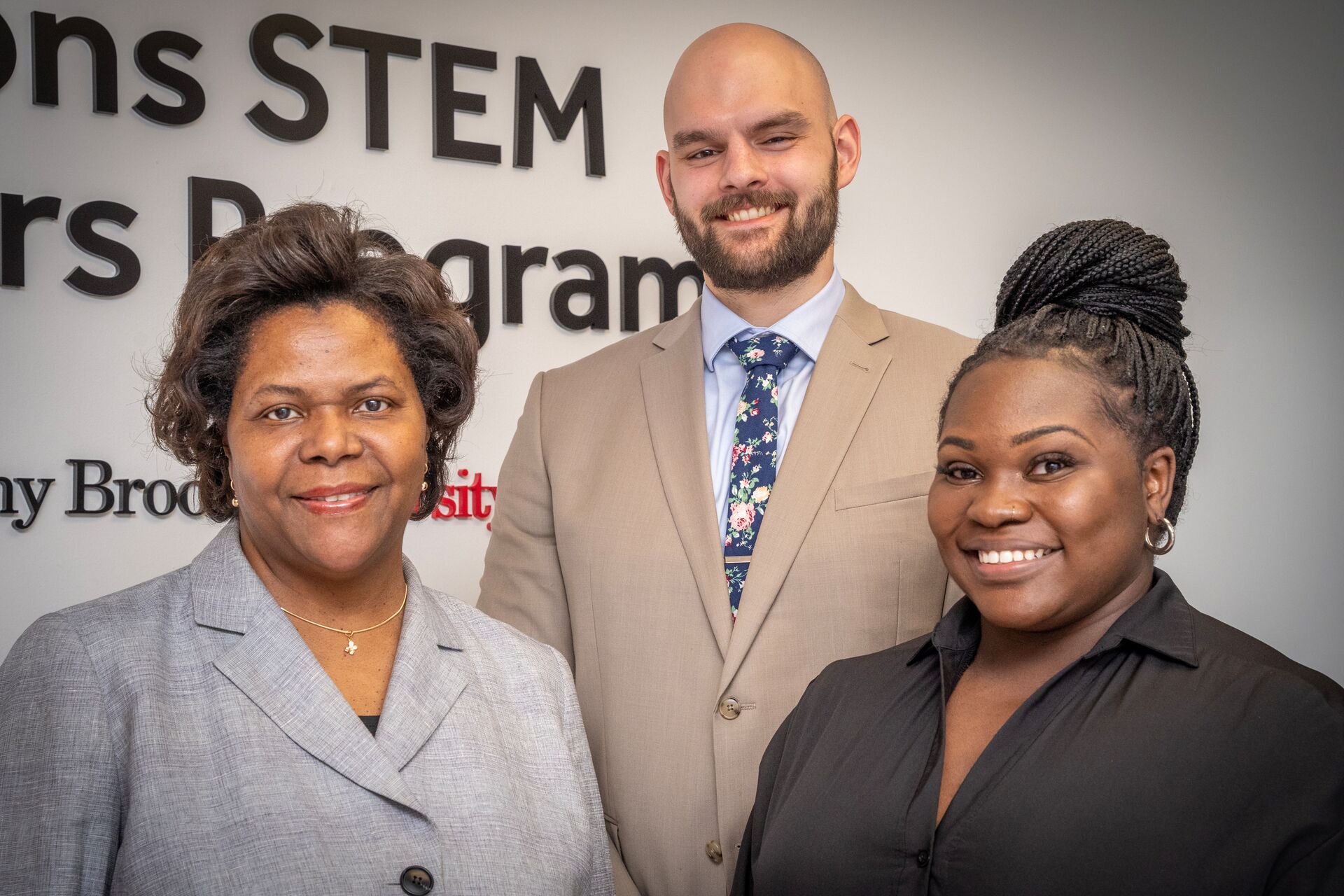 Three professionals smiling in front of a Stony Brook University "STEM Scholars Program" wall graphic.